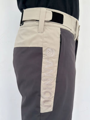 Close-up of a person wearing color-blocked ski pants with a beige upper section, dark gray lower section, and a vertical beige panel on the leg featuring the word 'ANNIOU' in raised embroidery.