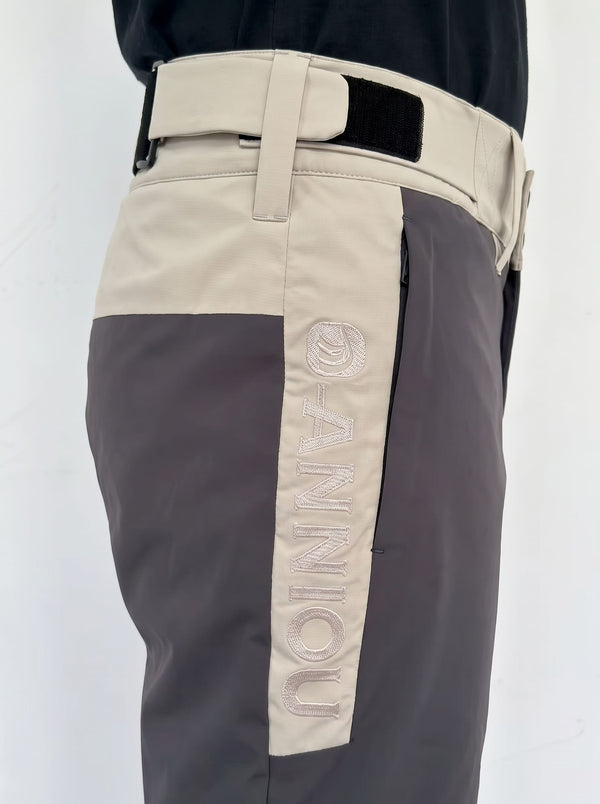 Close-up of a person wearing color-blocked ski pants with a beige upper section, dark gray lower section, and a vertical beige panel on the leg featuring the word 'ANNIOU' in raised embroidery.