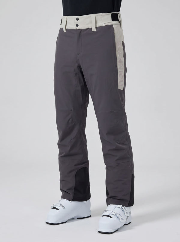 Dark gray ski pants with beige side panels and a matching beige waistband, shown on a model wearing white ski boots against a light gray studio background.