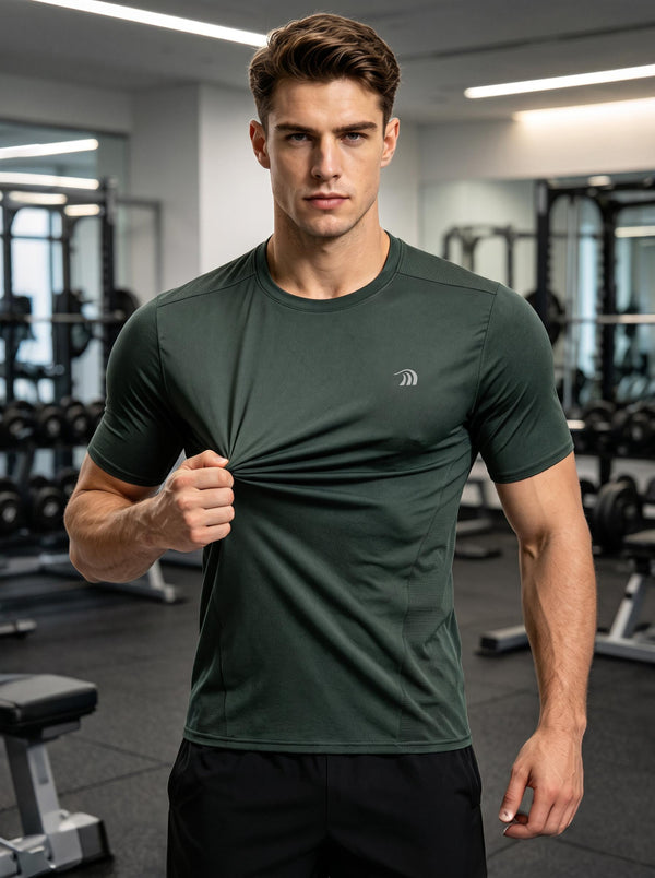 A man in a dark green athletic T-shirt stands in a gym, pulling at the fabric with one hand to show its stretch and texture, with gym equipment in the background