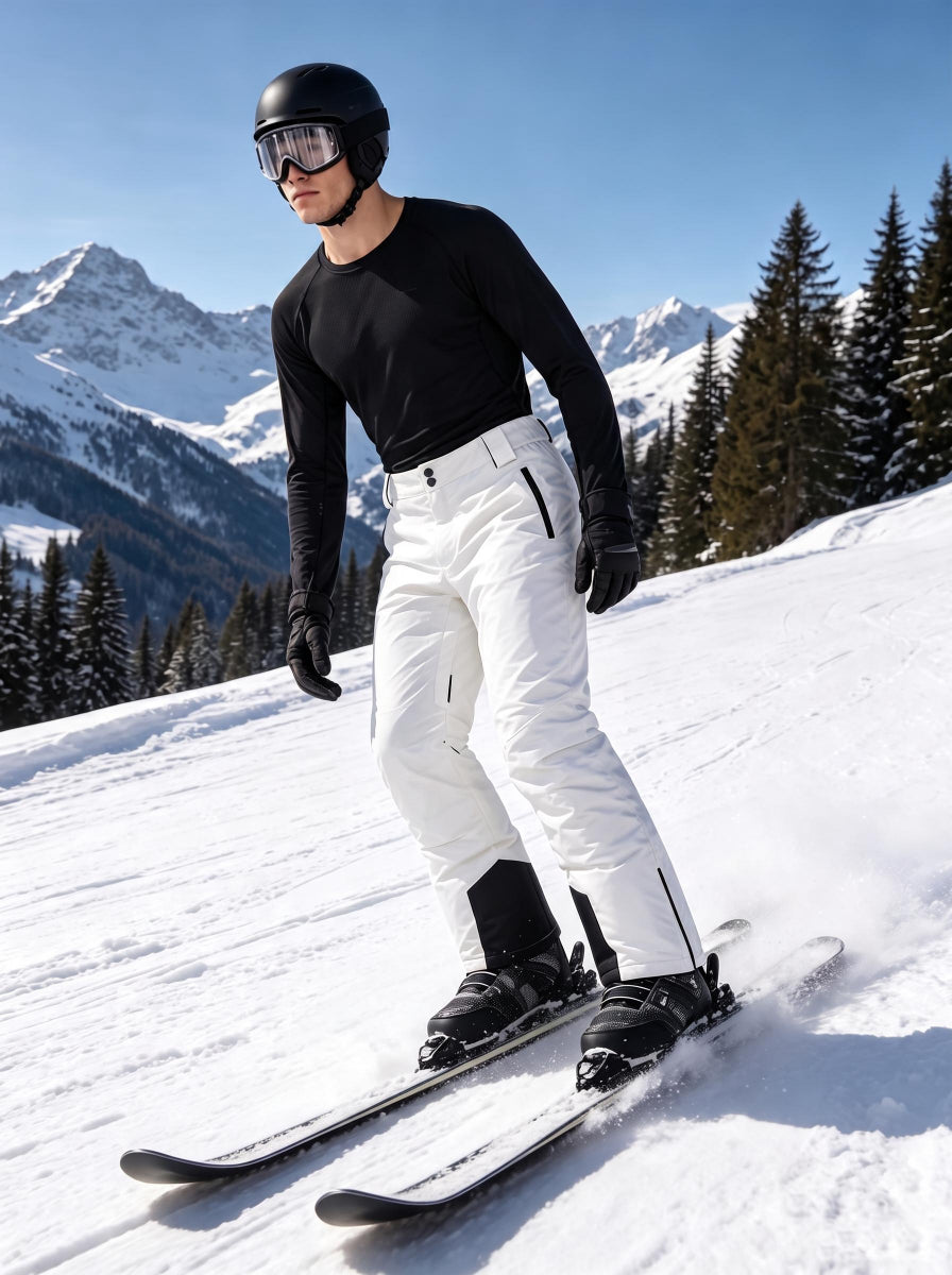 A male skier glides down a sunlit alpine ski slope, equipped with a black ski helmet, goggles and insulated ski gloves. He wears a black long-sleeve thermal top paired with crisp white insulated ski trousers featuring black zippered side pockets and reinforced abrasion-resistant black ankle panels. Classic snow-capped mountain ranges and snow-covered pine forests stretch across the background, with fresh snow spraying around his skis to highlight dynamic downhill skiing movement under clear bright blue skie