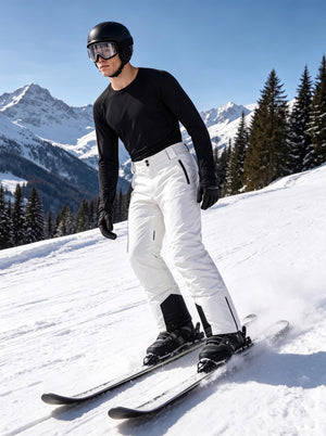 A male skier glides down a sunlit alpine ski slope, equipped with a black ski helmet, goggles and insulated ski gloves. He wears a black long-sleeve thermal top paired with crisp white insulated ski trousers featuring black zippered side pockets and reinforced abrasion-resistant black ankle panels. Classic snow-capped mountain ranges and snow-covered pine forests stretch across the background, with fresh snow spraying around his skis to highlight dynamic downhill skiing movement under clear bright blue skie