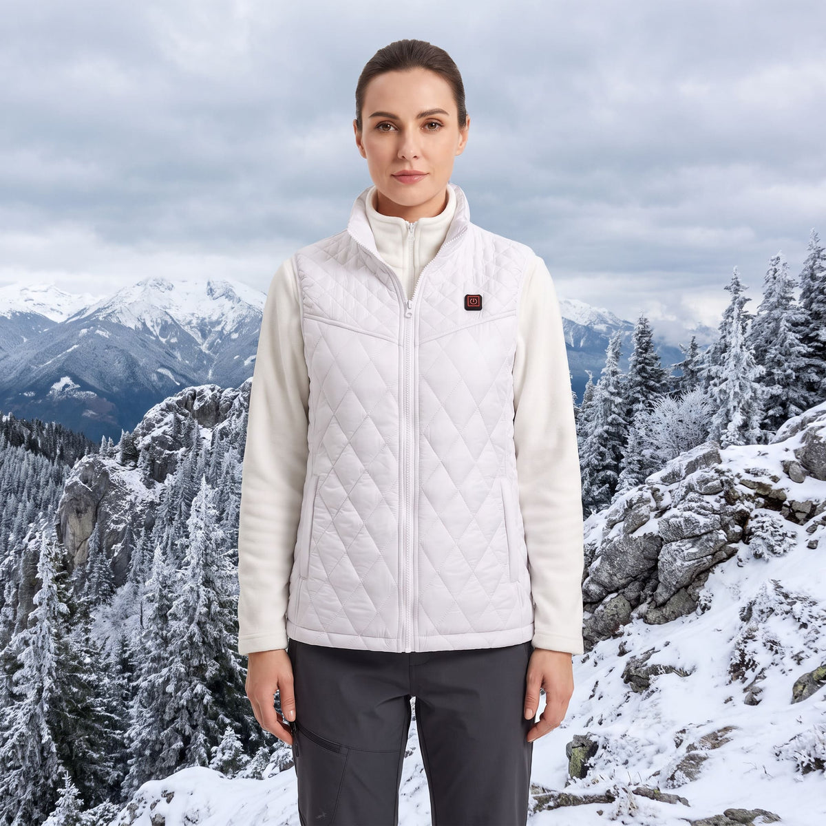 A woman stands outdoors in a snowy mountain landscape wearing a white quilted zip-front vest over a cream fleece jacket and dark gray pants.