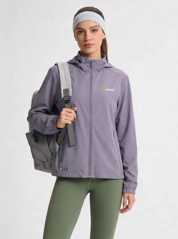 A woman modeling a #purple hooded athletic jacket with a green and white Anniou logo on the left chest, paired with olive green leggings and a gray backpack against a light gray studio background.