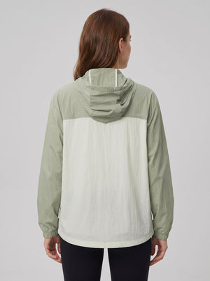 A woman viewed from the back wearing a lightweight hooded jacket with a sage green upper section and sleeves, and an off-white lower back panel, against a plain light gray studio background.