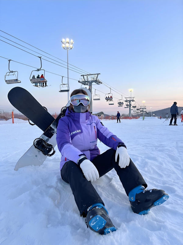 A young lady sits on the snow at a ski resort. She wears a gradient #purple ski jacket, black snow pants, white gloves, reflective ski goggles and a face covering. A black-and-white snowboard leans behind her. Ski lifts, tall floodlights and soft sunset twilight fill the background, with other skiers scattered on the snowy slope.