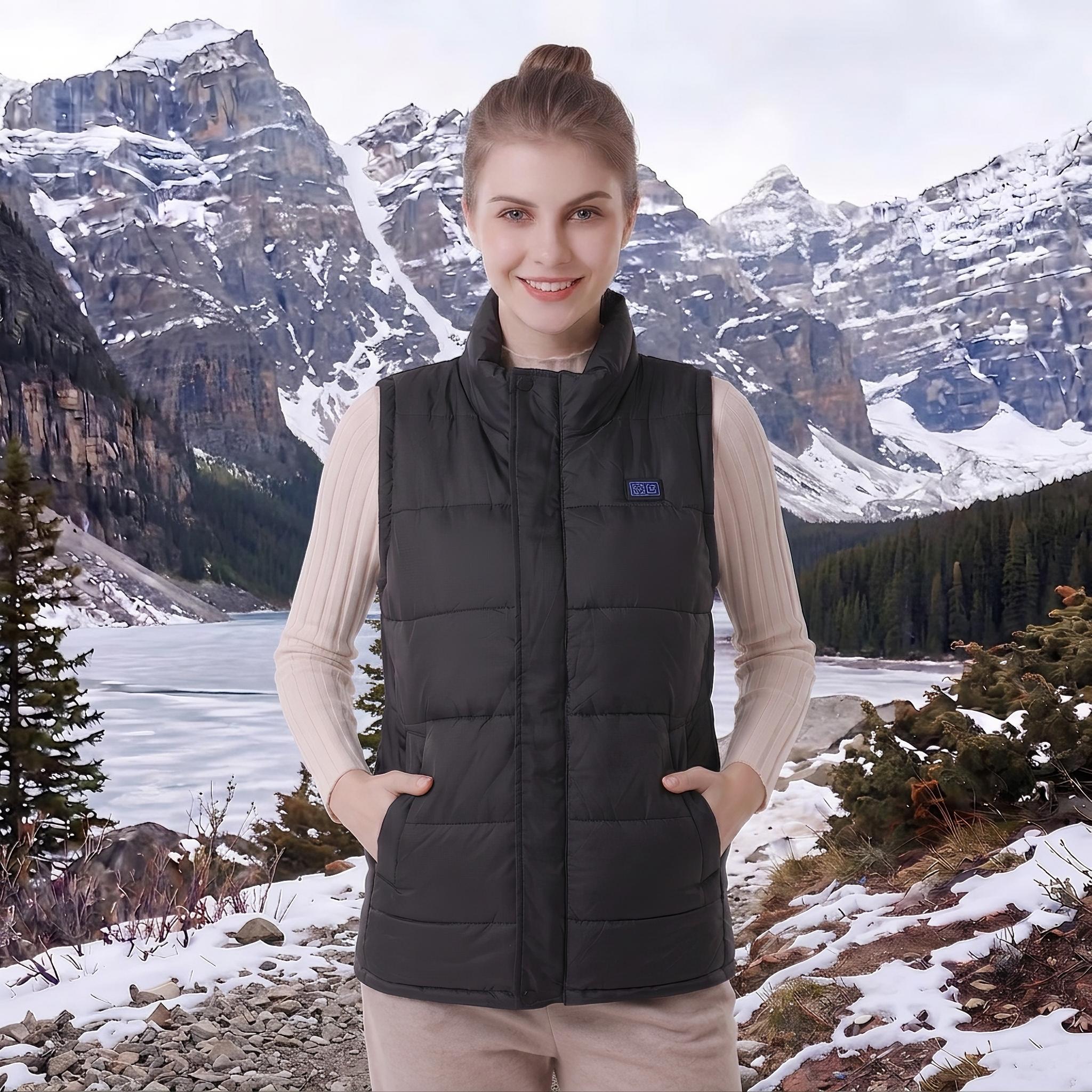 young woman with hair bun wearing black padded heated vest, hands in side pockets, standing outdoors against snowy mountain‑lake winter landscape, clear product display of women’s thermal gilet.