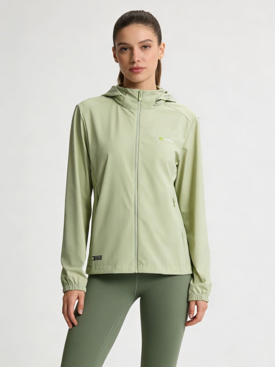 A woman modeling a #lightgreen hooded athletic jacket with a full zipper and matching sage green leggings against a plain light gray studio background.